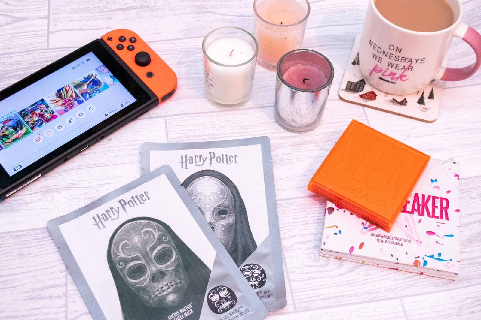 A flat-lay including a Nintendo Switch to the top right, two Harry Potter themed face masks at the bottom stacked, three votive candles in pink, white and lilac, a cup of tea in a white and pink mug on the top right and two eye-shadow palettes on the bottom left stacked one on top of the other.