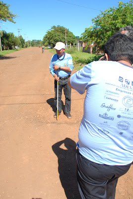 De costas para a foto está Marcos usando a camiseta da ADEVASB e com a câmera nas mãos, ao fundo está Luiz segurando a bengala e em cima de um buraco no meio da rua de terra.