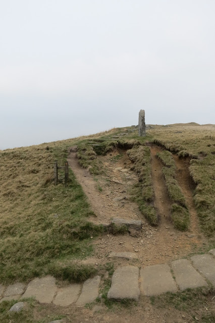 A stone laid path cuts across the foreground and a path of bare earth leads uphill at 90 degrees from it. A stone post acts as a waymarker for the second path.