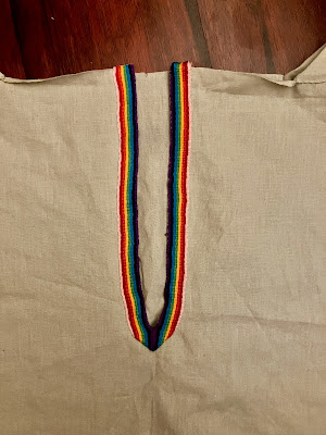 A close up of the neck slit of a square-cut shirt being constructed out of a light sage green fabric. The neck slit has been finished to the outside with a ribbon woven in the colours of the original gay pride flag. The top of the neck hole is still raw, with no overlocking. End ID.