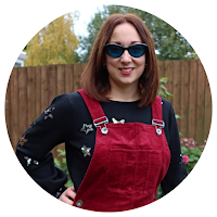 A caucasian woman with dark hear and tinted glasses wearing a jumper and red pinafore dress.