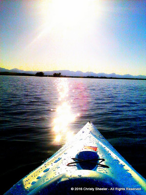 Sun glistens off the water's surface and the nose of the blue kayak.  The mountains all pale blue gray are at the horizon.