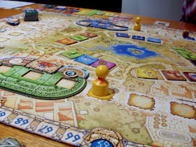A view of a game of Colosseum in progress. On the outer edge of the board is the scoring track, running all the way around the edge. Inside of that is the movement track, a series of square spaces with a bit of room between them. Two yellow pawns, representing Senators, can be seen on these spaces. Overlaid on this track are two colosseums, one blue and one green, both made up of two rounded end pieces and a squared-off middle piece. These are both laid upon the track so that the spaces in the middle of each piece aligns with a space on the track beneath. In the centre of the board is the marketplace, a group of five groups of three square spaces. Each space in the marketplace is occupied by a tile representing one of the elements that can be used to create your show: chariots, horses, poets, and so forth.