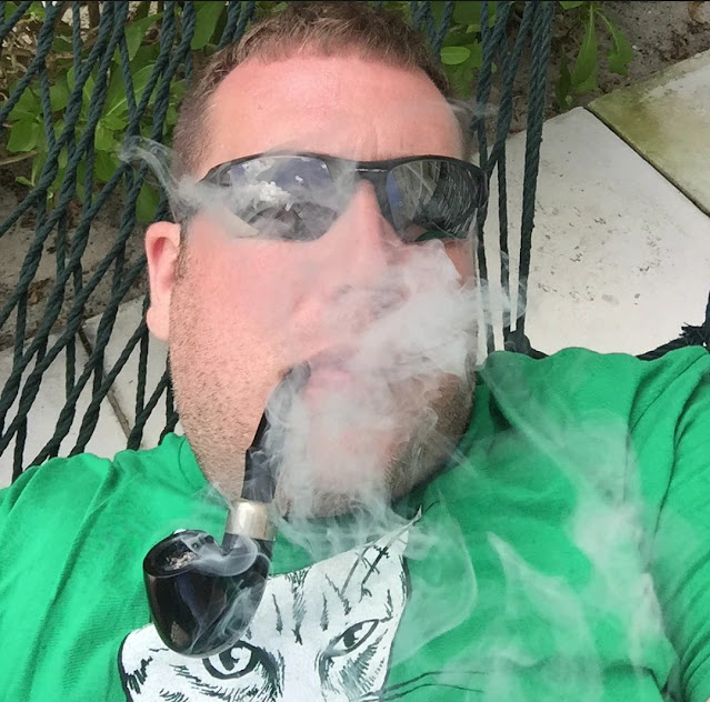 From the chest up wearing sunglasses and a green t-shirt smoking a pipe