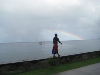 An Islander walking on a concrete wall next to the ocean with a rainbow above his head