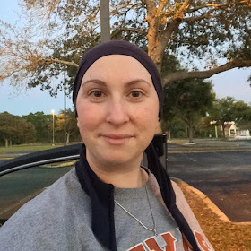 A woman wearing a head covering and sweatshirt in a parking lot with trees in the background