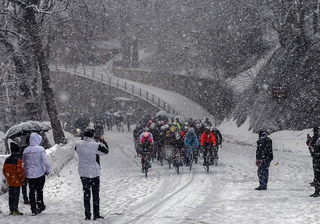 ciclistas competindo numa pista nevada. Nos arredores da pista, sarapintada por árvores temperadas, alguns observadores assistem a competição.