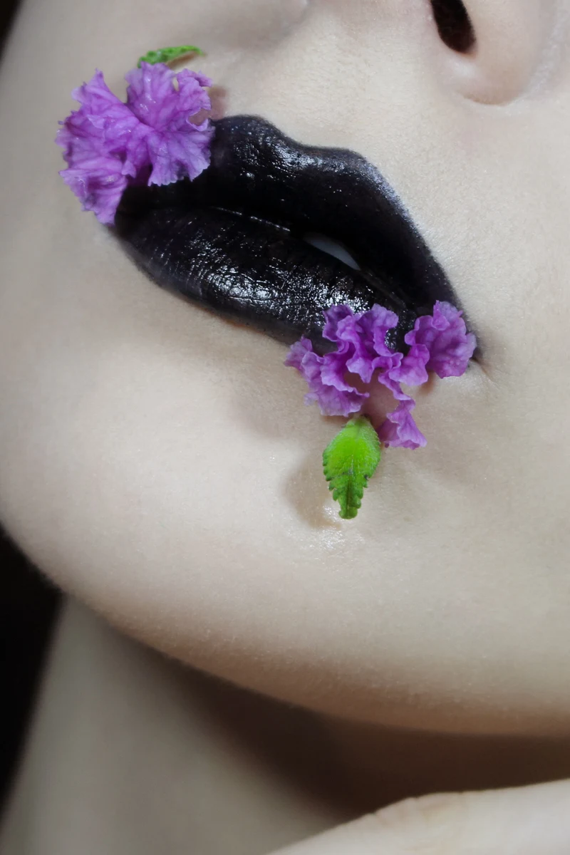 close-up of woman lips with editorial black lip look decorated with fresh purple flowers