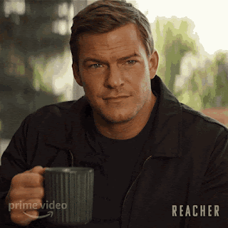 Alan Ritchson as Reacher is sitting outside, lifting a mug to his mouth. He is a big guy. His short hair is brushed neatly and he has some facial scars from a recent fight. He looks calm yet quizzical as he sips.