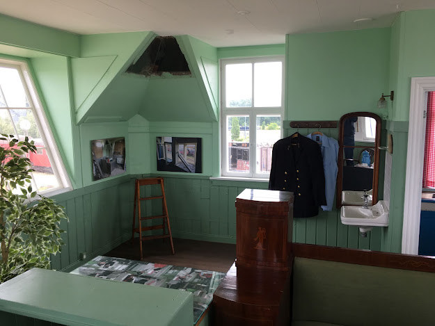 An hellgrünen Wänden hängen Bilder und Uniformen. Ein kleines Waschbecken in der Ecke. Davor ein kleiner Holzschrank.