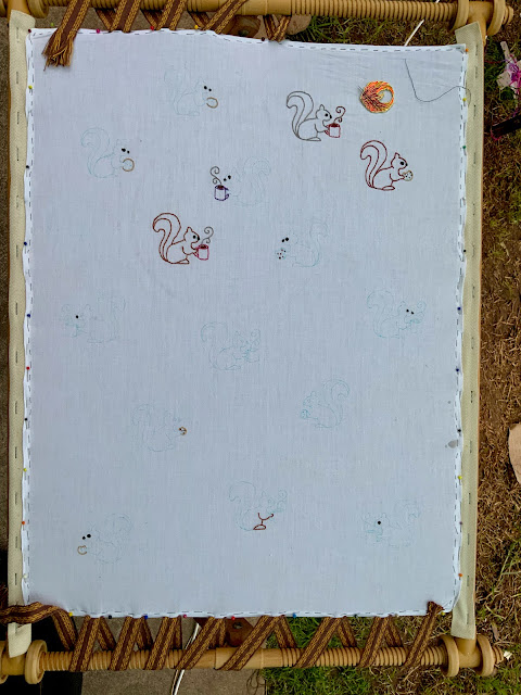 A large piece of white cotton fabric is stretched in a tapestry frame, held in place with sewing pins on the long edges and pinned, woven inkle bands on the short edges. There are 15 outlines of cartoon squirrels marked on the fabric in blue, water-erasable ink. Each squirrel is holding either a mug or a cookie. Three of the squirrels have been completely embroidered in split stitch. Several more have their noses and eyes embroidered. There is a needle minder in the top right corner in the shape of a phoenix. End ID.