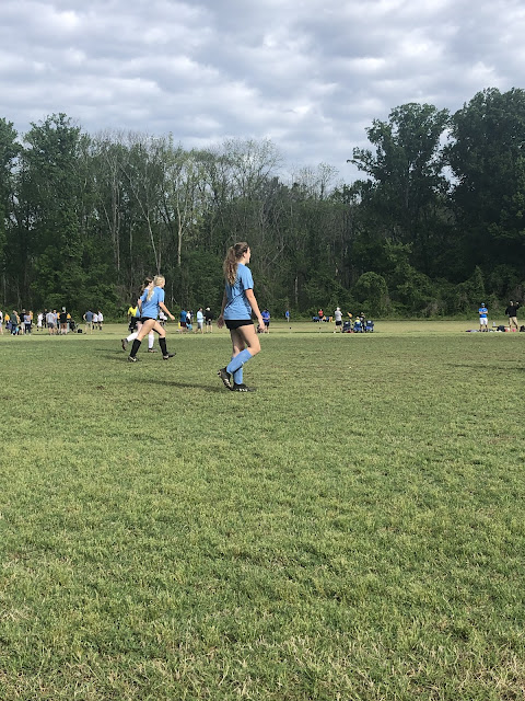 Elizabeth watching the ball on the opposite end of the field. She has her hair in a ponytail and is wearing her baby blue soccer jersey with matching socks, black shorts and her cleats.