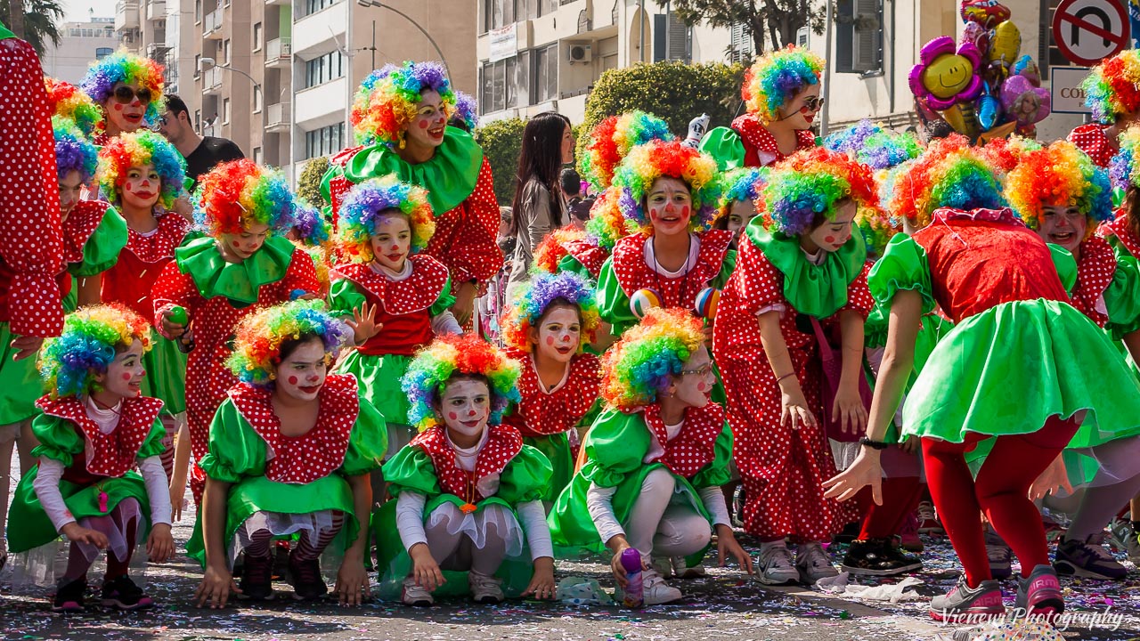 Wesoła i bardzo kolorowa grupa dzieci przebranych za klowny podczas dziecięcej parady karnawałowej w Limassol w 2014 roku.  Dzieci są naśladuja nauczycielke pokazującą. Zaczynają z przykucu.