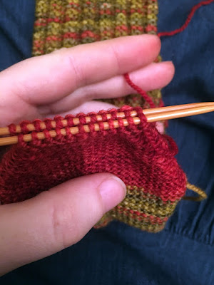 A close-up of two rows of knit stitches held on parallel wooden needles at the red toe of a sock, held up by a white hand.