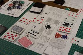 A table with assorted playing components, dice and playing cards, and play sheets and mats.