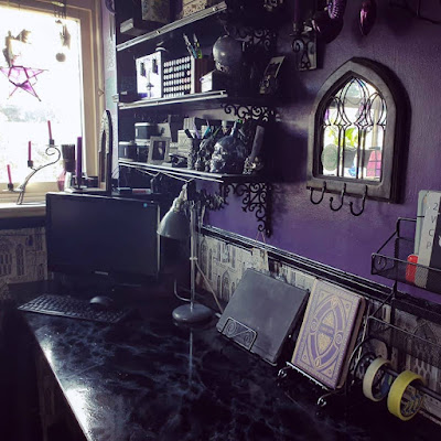 A photograph looking along a faux-marble desk towards a window and corner. The window is so bright that it looks mostly white, with a purple pentagram lantern barely visible. There is a curly black wrought iron candle-stick on the window-ledge, with three small purple candles in sockets each slightly higher than the one before, and the purple candles are at slightly jaunty angles. There is a small purple vase on the window-ledge with incense sticks. Several wind-chimes hang in the window, indistinct in the bright light. The window-frame is off-white. Black moulded dado-rail runs from under the window-ledge and around to the adjoining wall. On the adjoining wall, into the corner, there are a set of shelves, three of which are visible; they are above the desk and dado-rail. The shelves are black on ornate Gothic style reproduction Victorian cast-iron brackets, and the front edges of the shelves have thin moulded trim along them. On the shelves are an assortment of tubs and boxes, ornate with predominantly black, grey, silver and purple as the colours. There are also several skull ornaments, and a lavender glasss orb hanging from the second bracket up, nearest the camera. In the corner is a black Lenovo computer, turned off; the monitor is obscuring most of the PC tower, and the keyboard and mouse are visible; the mouse has been left on the keyboard. The faux marble desk is mostly clear except a grey vintage-style desk-lamp, a tablet computer resting on a black swirly metal recipe book stand, and an object that looks like a book with an ornate purple and gold cover, but which is actually a Harry Potter theme lamp with the cover reading 'Liber Lux'; the Harry Potter book-lamp is on a silver recipe-book stand. The wall adjacent to the window, above the desk, is mostly a dark, rich purple. On the purle wall, nearer the camera, there is a mirror in the shape of a wide Gothic arch in a wooden frame; the mirror glass is subdivided horizontally into three thin Gothic arches, two shortm one tall one in the middle - with wire. At the bottom of the wooden mirror frame are three black metal hooks unused. Above the mirror, three purple glass hearts - two bright purple, almost fuchsia and one dark violet - are visible suspended, but what they are suspended from is cropped off at the top of the photograph. Under the dado rail that runs from under the window-ledge along both visible walls is a greyscale wallpaper of Gothic arches, a reproduction if an 1830's style, with a slightly crude wood-block effect. On top of the PC tower in the corner is a mostly wooden candle-holder; it is an ornately carved wooden Gothic arch with a mirror in, on a wooden base, with a purple taper-candle in a brass socket in front of the mirror; the candle-holder is at an oblique angle. Also in the foreground is a metal spice rack, in the same twisted metal tubing and spiral design style as the recipe-book stands; it is slightly thicker metal and powder-coated matte black. At the bottom of the spice-rack is a spindle supporting two visible rolls of tape, on the top shelf of the spice-rack, cropped from view, are art materials in shallow metal tray-tins. The re-purposed spice rack is in the fore-ground, at the far right of the image, and is partially cropped. The image is poorly lit as the room is dingy, and the window is much, much brighter than the room; the effect is dark, subdued and closed-in. The whole collection of visible objects gives a witchy, eclectic aesthetic, especially the purple walls and ornament on much that is visible. The photograph looks like it was taken in the afternoon.