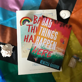 A photo of a Bad Things Happen Here by Rebecca Barrow proof on a Progress Pride flag over the POC chevron, which is at a diagonal, so the chevron is pointing to the bottom right, On top of the proof, on it's right side, is a postcard of the book's final cover - which is different to the proof cover - on a diagonal, top right, to bottom left. There is a rainbow pin on the top left of the proof, and around the book is a white shell, a shiva shell heart, and a small jar of sand and tiny shells.