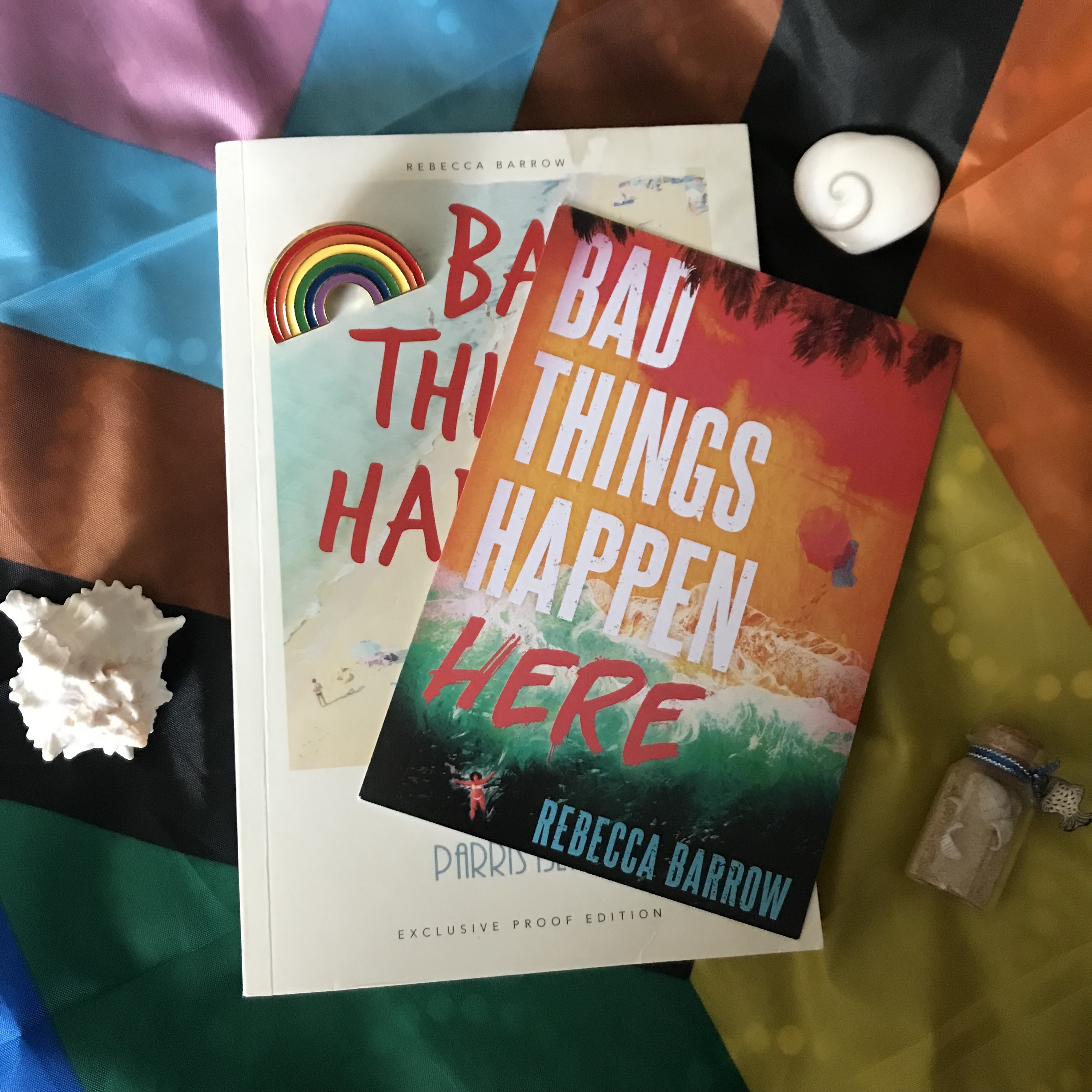 A photo of a Bad Things Happen Here by Rebecca Barrow proof on a Progress Pride flag over the POC chevron, which is at a diagonal, so the chevron is pointing to the bottom right, On top of the proof, on it's right side, is a postcard of the book's final cover - which is different to the proof cover - on a diagonal, top right, to bottom left. There is a rainbow pin on the top left of the proof, and around the book is a white shell, a shiva shell heart, and a small jar of sand and tiny shells.