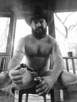 Black and white big huge bear of a cowboy shirtless wearing jeans and holding a cigar towards the camera