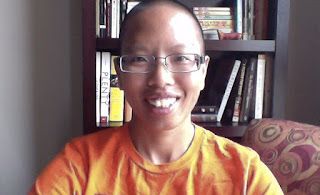 a close headshot of liz facing the camera directly; she is in front of a bookcase wearing an orange shirt; she smiles with the light coming from her left