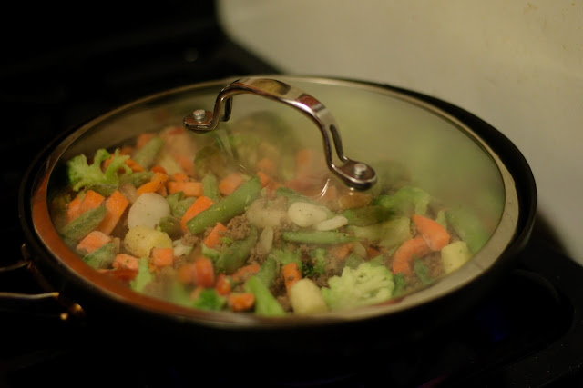 A lid on the pan with the ground beef and the frozen vegetables on the stove.