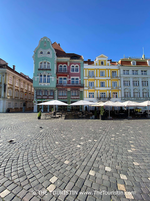 A sandstone tiled square with a cafe in front of a row of period houses with pink and light green painted facades, ornate roofs, Secession-style decorations, and ceramic details under a bright blue sky.