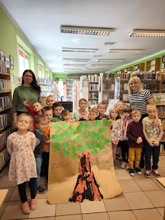 Zdjęcie grupowe pani bibliotekarki, pani przedszkolanki i grupy przedszkolnej. Wszyscy stoją przed ladą biblioteczną. W tle widać regały z książkami i wystawkę książek o drzewach. Kilkoro dzieci stojących pośrodku trzyma w rękach wykonaną wspólnie pracę plastyczną rysunek dużego drzewa.