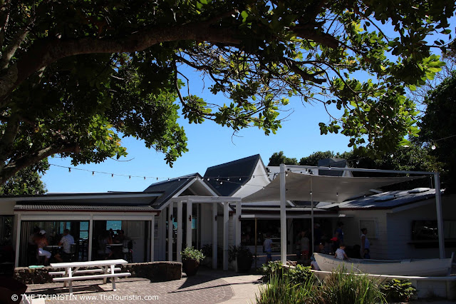 Terrace with a rowing boat in front of a large grey weatherboard house and a glimpse of the ocean through the windows of the property.