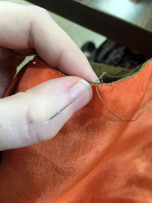 A close-up of a needle inserted through the edges of sheer peach and green-gold silk, turned in and whipstitched together. The needle also happens to be inserted through a layer of the skin of the thumb it's skimming along, where a white hand is pinching the edges of the fabrics.