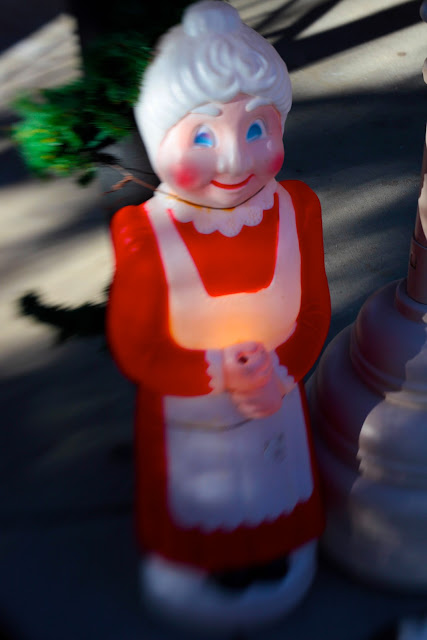 A strange Christmas and a smiling Mrs. Claus lawn ornament tied to a post by her neck. 