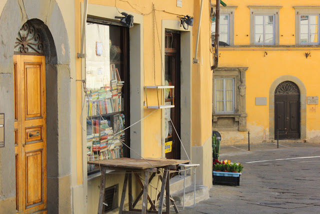 Na rogu niewielkiego Piazza Gamurrini w Monte San Savino znajduje się księgarnia. Przed sklep wystawione są puste jeszcze stoły (jest wcześnie i sklepik jest zamknięty). Witryna księgarni zastawiona jest szczelnie ułożonymi na półkach książkami. Żółć fasad miesza się z szarością bruku i kamiennych ościeży drzwi.