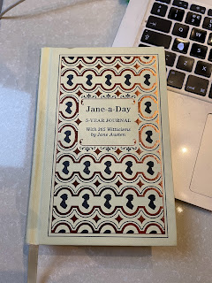 A small, yellow notebook. It has an embossed gold pattern over the front. The title says, 'Jane-a-Day' and underneath in smaller writing, it says, '5-Year Diary. Beneath that, it says, 'With 365 Witticisms by Jane Austen.'