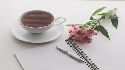 White top online work table with pen and notepad, a mug of coffee and pink flowers.