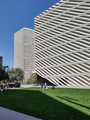 Photo of two buildings, with a grassy area in front of them and blue skys behind them. The far building is  maybe 15 or 20 stories, with a concrete window grid. The building in the foreground is four storeys with repeating concrete blocks with diagonal apertures in the facade. It's the Broad Museum, on Grand in downtown LA.