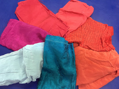 Seven folded remnants of silk fabric arranged in an overlapping spiral. Half are bright, saturated oranges and reds, one is screaming magenta, one pale robin's egg blue, and one clear turquoise, on a cobalt blue background.