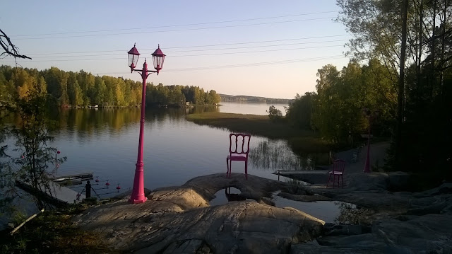 Kirkonvarkauden järvi on täysin sulanut, puut vihertävät. Kalliolla on punainen katulamppu sekä punainen tuoli kohdistettuna vastarannan horisonttiin.