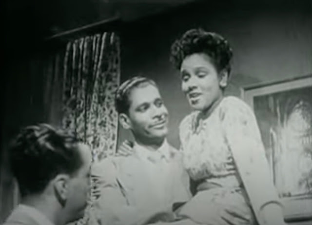 Screenshot from the 1947 movie "Boy! What a Girl" where Ann Cornell is sitting on top of an upright piano and one of the other musicians is smiling while holding her and watching her sing.