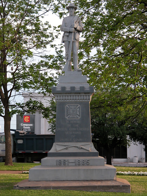 This photo shows the stone statue of a man in a Confederate uniform, standing and holding a rifle atop a base that reads, “In honor of the United Confederate Veterans of Parker County, 1861-1865.” The base was placed on the grounds of the Parker County Texas Courthouse by the United Daughters of the Confederacy in 1915, but the statue’s date and ownership are less clear.