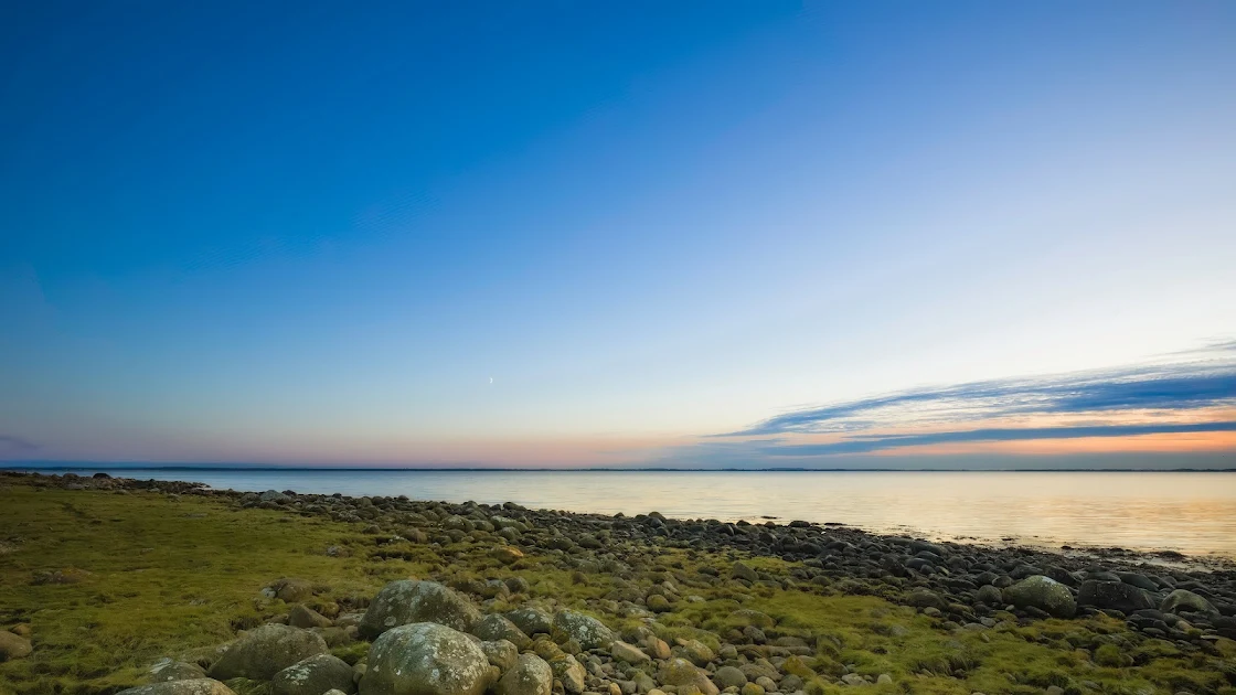 4K image featuring a gentle sunrise over a calm sea with a rocky shoreline under a vast blue sky.