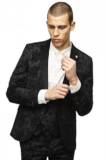 A man in a dark purple-black suit with black floral setail all over with a bright white shirt on a white background. 