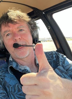Sexbot author Patrick Quinlan in a blue shirt and headset, giving the thumbs up in a helicopter cockpit near Sarasota Florida