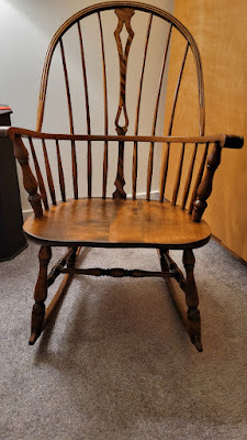 Photo of a wooden rocking chair with a reddish wood stain sitting on beige carpet in the glow of a small lamp. The back of the rocker has rounded spindles, with the one closest to the edge of the seat having more curves. A rocking chair collector described it as an army knuckle arm Windsor rocking chair with saddle joints where the legs meet the rockers.