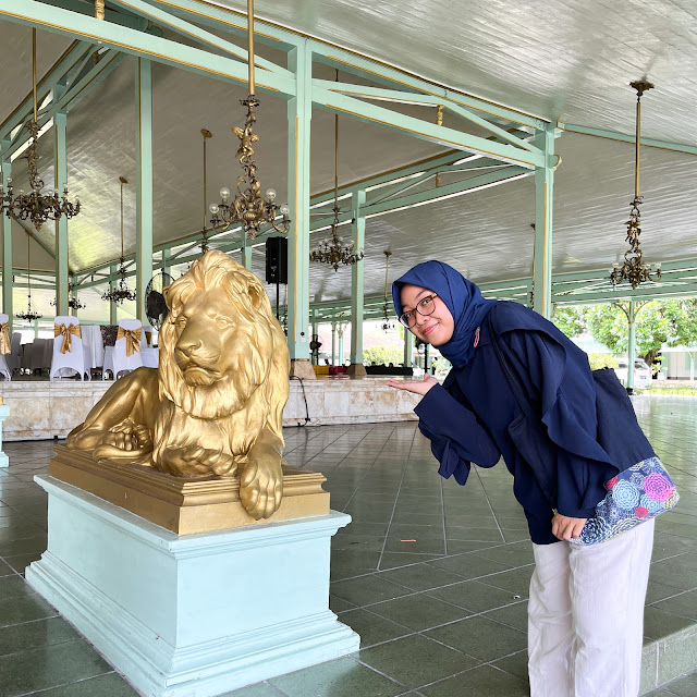 Seorang wanita alias aku berfoto di depan sebuah patung singa emas. Si singa emas sedang duduk selonjor. Wajahnya menghadap ke arah kiri.