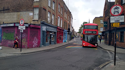 A narrow section of street with shops on both sides, narrow footways and the the restriction signs. There is a rainbow painted on the road and a bus coming towards us.