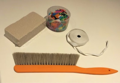 Shows a sponge, a plastic tub full of paper clips, a roll of ribbon and a brush