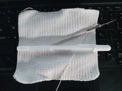 A crocheted rectangle lying on its side. The white thread it's made from is hanging out at the top left corner and from the middle of the right side. The steel hook is still attached, and there is a white gel pen laying across the middle horizontally for scale.