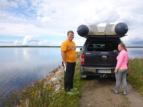 Kumivene maastoauton katolla, kaksi henkeä seisoo auton takana, auto ajettu kapealle niemekkeelle keskelle vettä