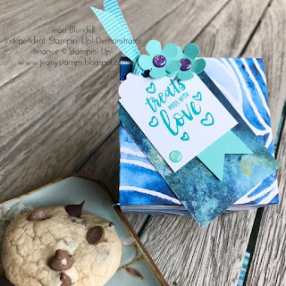 Square giftbox with blue and white swirly pattern and a gift tag saying "Treats made with love", decorated with small punched flowers and gems. The box is sitting next to a small turquoise plate with a chocolate chip cookie on it.