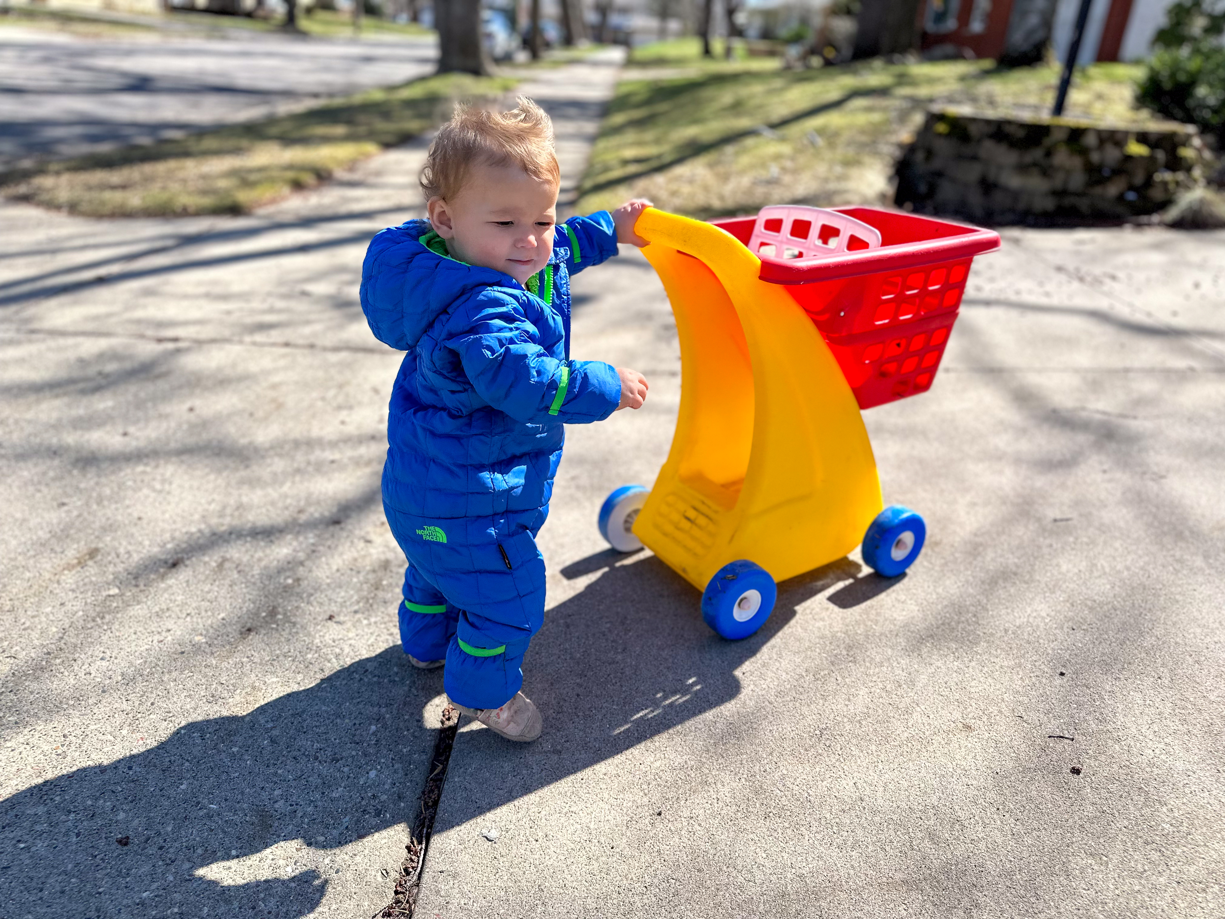 13 month old Montessori Toddler pushes a toy shopping cart while outside