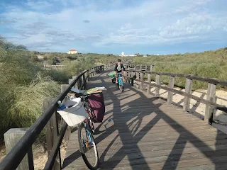 Boardwalk with railings, in front my bicycle, in the background Aim'jie riding, on the side sand and bushes.